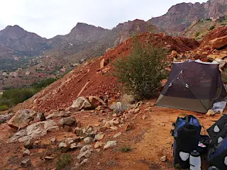 Zeltlager mit grauem Zelt und Ausrüstung auf rotem Erdhang vor bergigem Panorama im Antiatlas