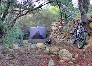 Zelt steht auf unebenem Boden in bewaldeter Berglandschaft neben einer Steinmauer, Fahrrad lehnt an Baum