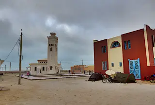 Dorfplatz in El Argoub mit einem Minarett, mehreren Zelten und einem großen Gebäude mit blauer Tür unter bewölktem Himmel