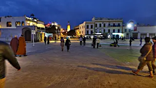 Abendliche Szene auf einem Platz in Essaouira mit mehreren Personen, umgeben von weiß getünchten Gebäuden und einem beleuchteten Turm