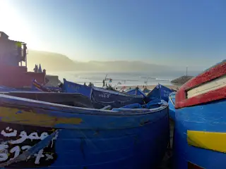 Mehrere blaue Fischerboote stehen dicht nebeneinander in der Bucht von Imsouane bei Sonnenaufgang mit Bergen im Hintergrund