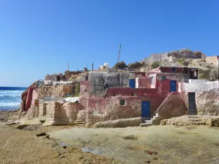 Mehrere einfache Hütten aus Stein und Lehm mit blauen Türen stehen am Strand von Imsouane unter klarem Himmel