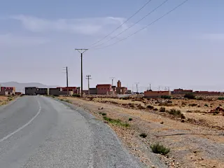 Landstraße mit Strommasten führt zu einem kleinen Dorf mit flachen Gebäuden in einer trockenen, kargen Landschaft