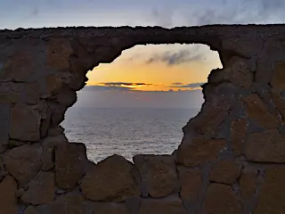 Durch eine unregelmäßig geformte Öffnung in einer Steinmauer ist das Meer mit einem Sonnenuntergang am Horizont zu sehen
