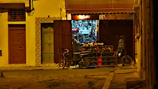 Zwei Personen stehen in einem kleinen, beleuchteten Laden mit Regalen voller Flaschen und Dosen, davor stehen zwei Motorräder 