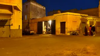 Straßenkreuzung in Trabsini bei Abenddämmerung mit gelb beleuchteten, Gebäuden und zwei Personen in traditioneller Kleidung