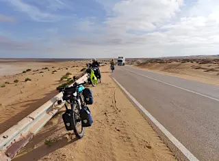 Zwei beladene Fahrräder stehen am Straßenrand einer Wüstenstraße, im Hintergrund fahren ein Reiseradler und ein Lastwagen
