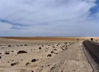 Asphaltierte Landstraße mit Verkehrsschild und Fahrzeug am Rand einer sandigen, kargen Wüstenlandschaft unter bewölktem Himmel