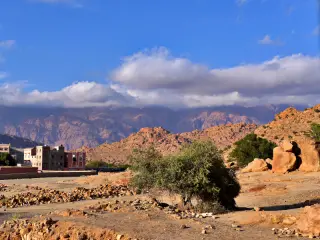 Tafraout mit Felsenlandschaft, einzelnen Gebäuden und einem Baum im Vordergrund unter blauem Himmel mit Wolken über den Bergen