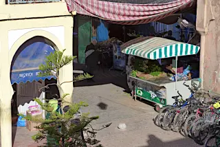 Gasse mit einem Marktstand unter einer gestreiften Markise, einem Laden mit blauem Vordach und einem Fahrradständer in Tiznit