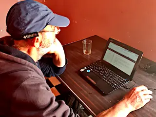 Mann mit blauer Kappe und Brille sitzt an einem dunklen Tisch und arbeitet an einem Laptop mit geöffnetem Textdokument