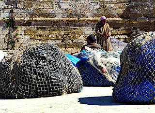 Zwei Fischer in traditioneller Kleidung reparieren Netze im Hafen von Essaouira vor einer alten Steinmauer