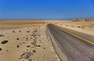 Asphaltierte Straße in der Westsahara mit gelber Mittellinie führt durch die Ebene einer Sabcha unter klarem blauem Himmel