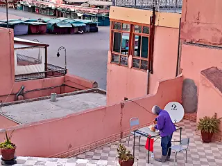 Person in Kapuzenjacke steht an einem Tisch auf einer Dachterrasse in Marrakesch mit zwei Stühlen und Pflanzen in Töpfen