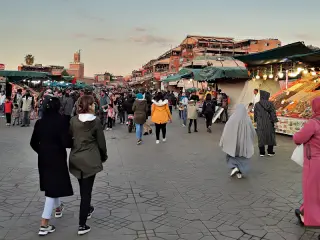 Menschen gehen über den belebten Platz Jemaa el-Fnaa in Marrakesch mit Marktständen und Gebäuden im Hintergrund