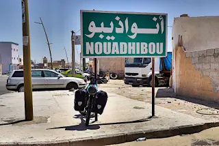 Ortseingangsschild mit der Aufschrift 'NOUADHIBOU' vor einer Straße mit parkenden Fahrzeugen und einem Motorrad in Mauretanien