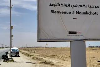 Werbeschild mit Begrüßung 'Bienvenue à Nouakchott' an einer staubigen Straße mit parkendem Auto und Fahrrad am Stadtrand