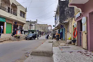 Straße in Dakar mit niedrigen Gebäuden, einigen Geschäften, Menschen und geparkten Fahrzeugen unter bewölktem Himmel