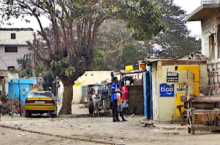 Straße in einer Vorstadt von Dakar mit großen Baum, mehreren Personen, einem Pferdewagen und gelben Taxi und kleinen Kiosk 