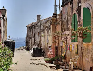 Enger Weg mit verwitterten Gebäuden und grünen Fensterläden auf der Insel Gorée mit Blick auf  die Skyline von Dakar 