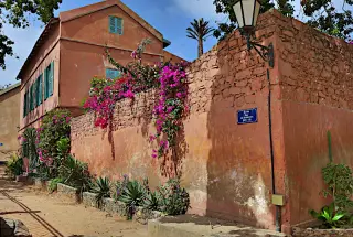Straßenecke auf der Insel Gorée mit hohen Mauerwerk, blühenden Pflanzen und einem zweistöckigen Haus mit grünen Fensterläden 