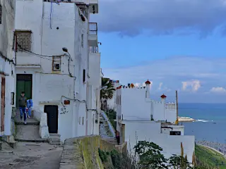 Weiße Gebäude säumen eine schmale Gasse an einem Hang mit Blick auf das Meer in Tanger, auf der zwei Personen  gehen. 