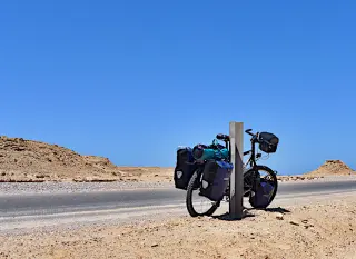 Ein mit Gepäcktaschen beladenes Fahrrad lehnt an einem Pfosten in einer Wüstenlandschaft unter blauem Himmel in der Westsahara
